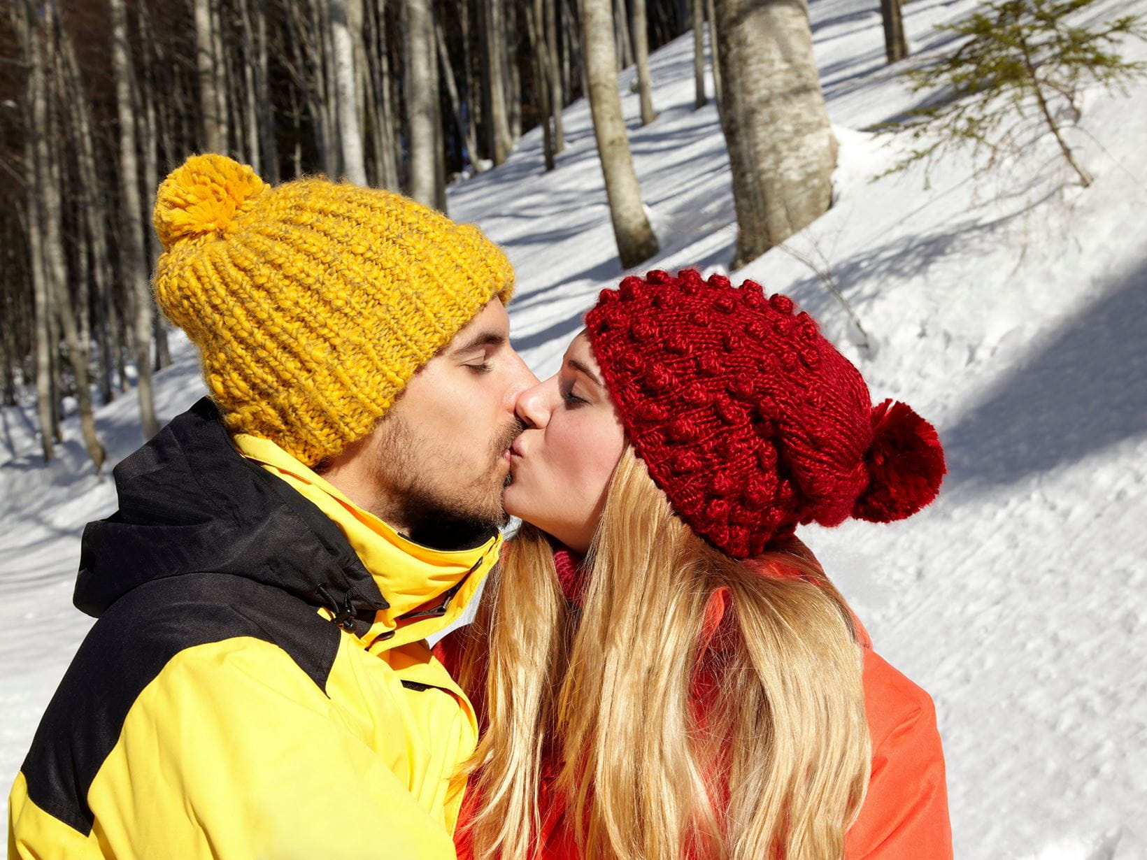 View of a male model wearing a yellow winter hat and jacket and a female model wearing a red winter hat and jacket kissing outdoors with a snow covered forest background.