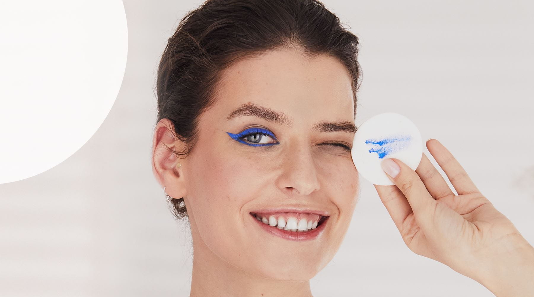 View of a female model wearing blue eyeliner holding a round cotton pad against a grey background.