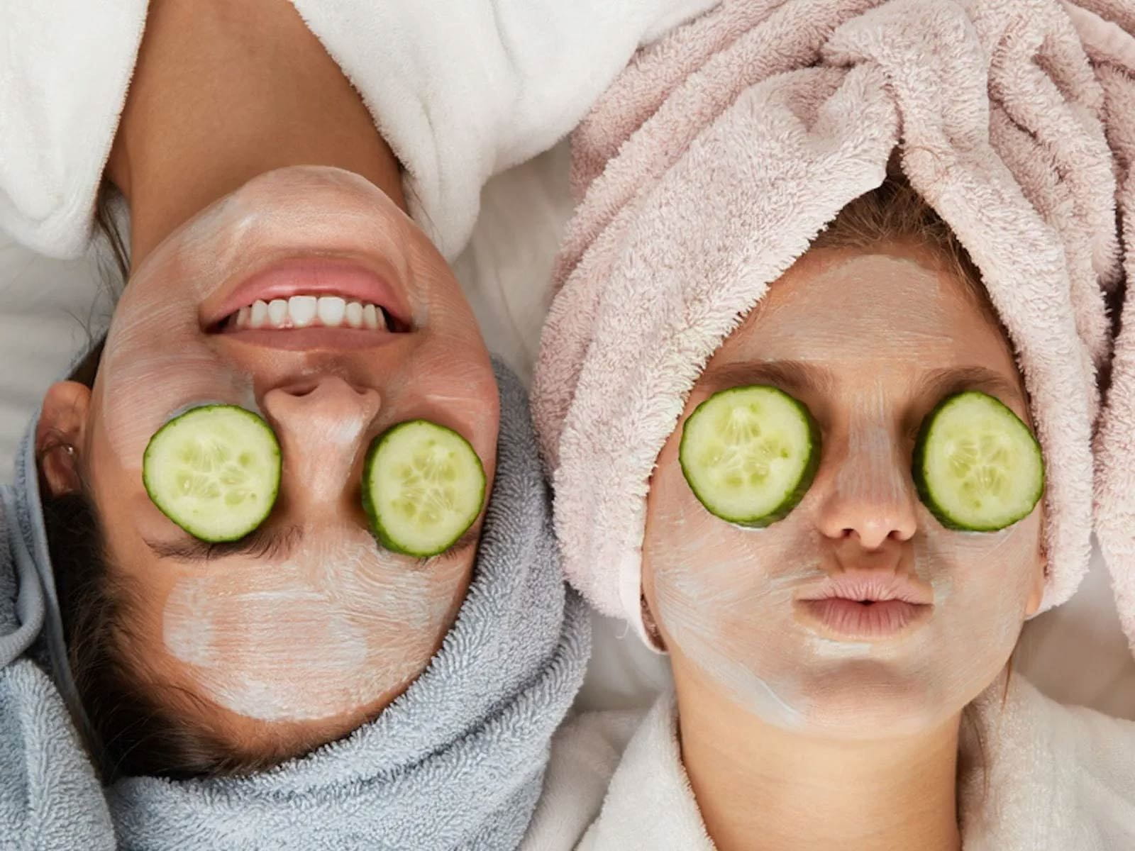View of two models with cucumber slices covering their eyes and towels over their hair while laying down side by side.
