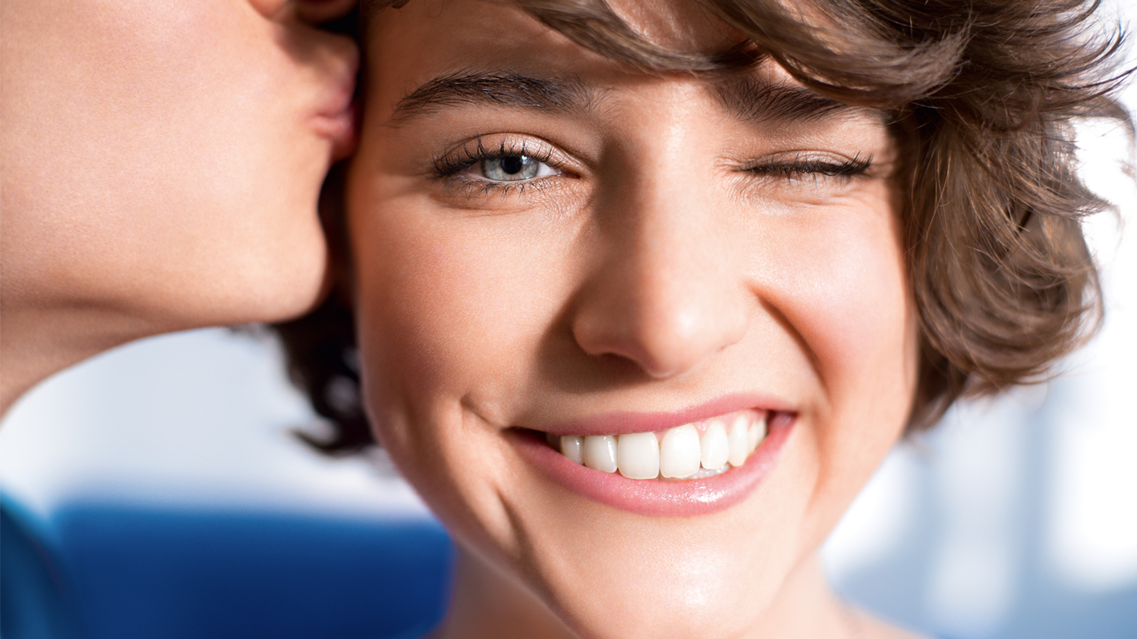 Smiling, brown-haired woman winking and being kissed on the temple.